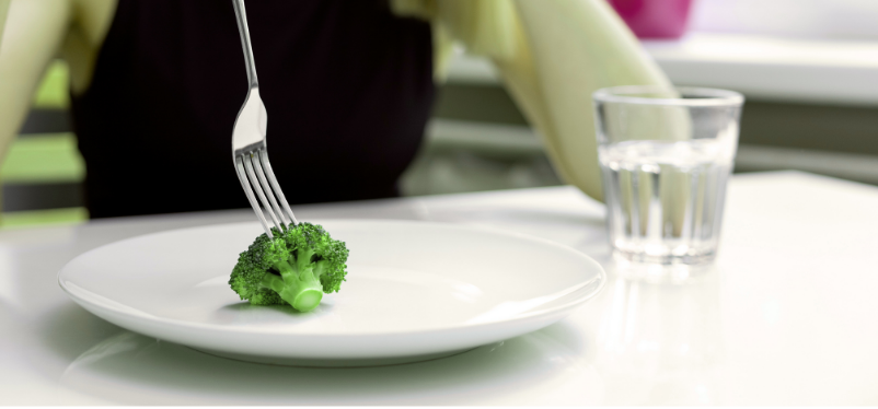 אישה יושבת ליד שולחן, בצלחת מונח ברוקולי אחד.