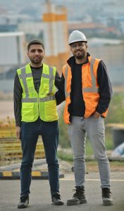 Two construction workers wearing safety vests and hard hats at a construction site.